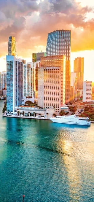 Coastal city skyline with tall buildings at sunset, turquoise waters, and cruise ship docked below golden sky