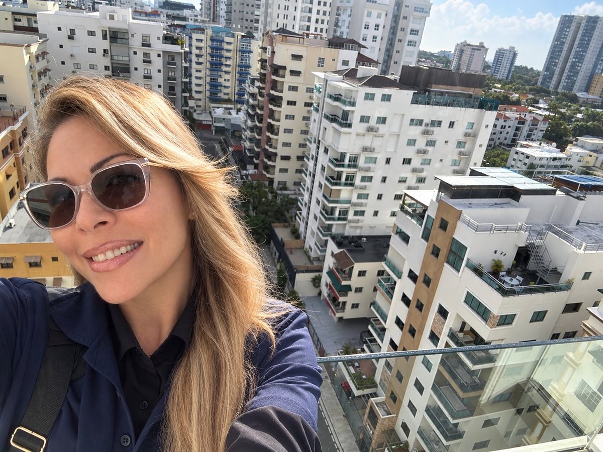 Woman with blonde hair and sunglasses taking a selfie on a high-rise building overlooking a dense urban cityscape
